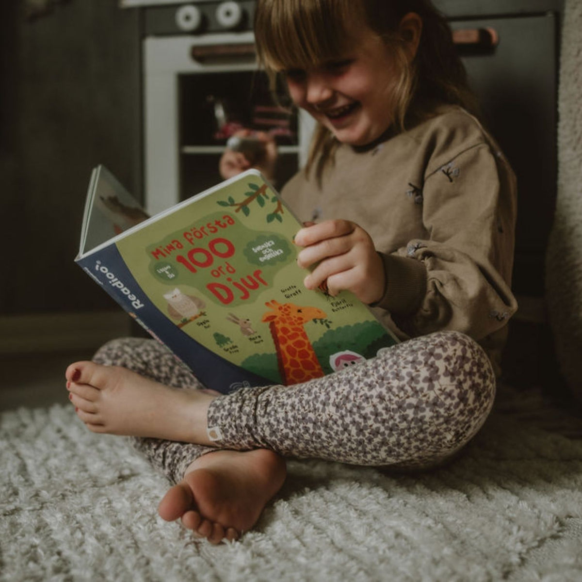 Flicka sitter på mattan och läser Readioo-boken "Mina första 100 ord: Djur".